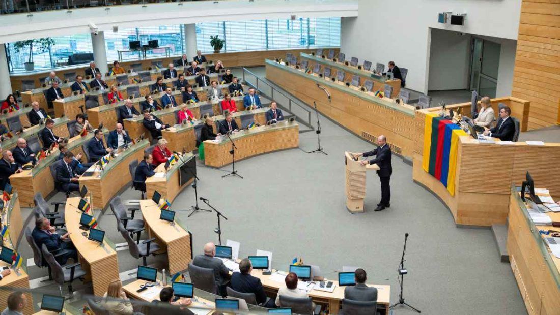 Žmonės sėdi parlamento salėje klausosi kalbančio vyro prie pulto
